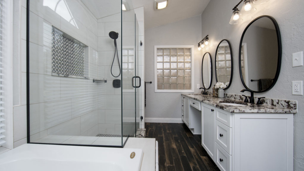 Modern farmhouse bathroom design featuring a glass-enclosed shower with custom tile, double vanity with round mirrors, granite countertop, and matte black fixtures, showcasing Marsh Cabinetry in a white finish.