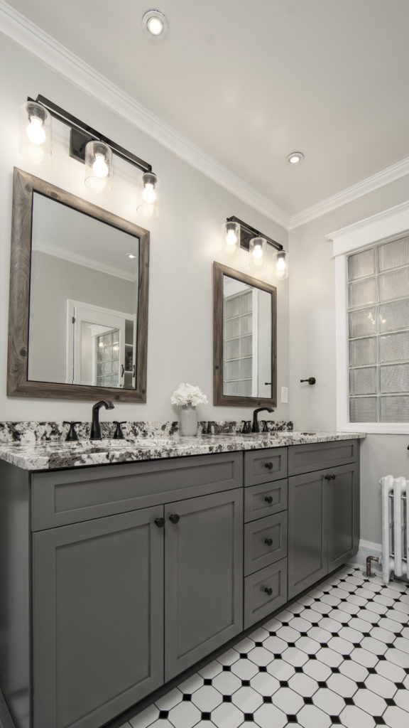 Modern farmhouse bathroom featuring Marsh Cabinetry in painted grey finish, double bowl vanity with granite countertop, matte black fixtures, rustic tile floor, and decorative mirrors.
